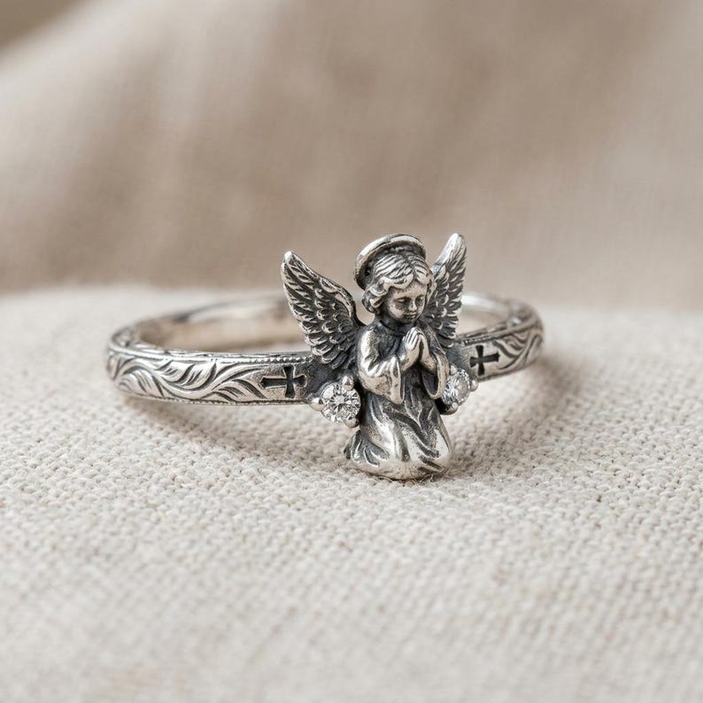May include: A silver ring featuring a three-dimensional kneeling angel with folded hands and wings. The band is decorated with engraved leaf patterns and small cross symbols, accented by two small clear round stones at the angel's base.