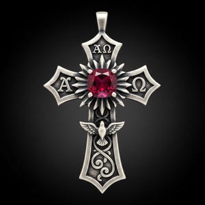 Può includere: Ciondolo a forma di croce argentata con una grande gemma rossa al centro. La croce presenta dettagli intricati, tra cui una colomba, elementi floreali e le lettere greche Alfa e Omega. Il ciondolo ha un'asola per una catena.