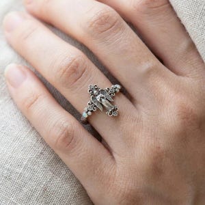 May include: A silver-toned ring featuring an intricate angel figure with outstretched wings as the centerpiece. The band is decorated with ornate scrollwork and small, iridescent opal-like stones embedded along the sides of the angel motif.