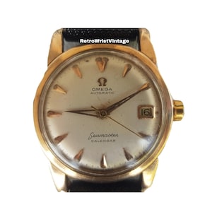 Könnte beinhalten: Vintage Omega Seamaster Calendar Uhr mit goldfarbenem Gehäuse und hellem Zifferblatt. Die Uhr hat eine Datumsanzeige bei 3 Uhr und goldfarbene Zeiger und Markierungen. Ein schwarzes Lederarmband ist sichtbar. Der Text "Omega Automatic Seamaster Calendar" ist auf dem Zifferblatt aufgedruckt.