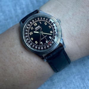 Puede incluir: Un reloj de pulsera automático Oris con esfera negra, caja de acero inoxidable y correa de cuero negro. La esfera del reloj presenta números blancos, un indicador de fecha y el texto "ORIS AUTOMATIC". El reloj se muestra en una muñeca.