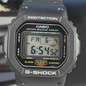 May include: A gray G-Shock digital watch with a black digital display showing the time and date. The watch face includes the Casio logo, and the text "WATER 200M RESIST". The watch has buttons for adjusting the time and light.