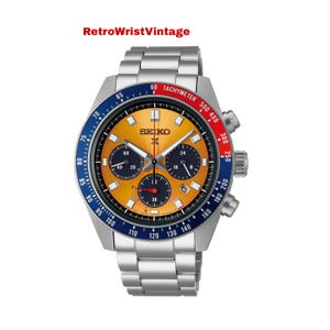 May include: A silver-toned Seiko chronograph watch with a yellow face, blue and red accents, and a stainless steel band. The watch face features multiple sub-dials and the text "SEIKO". The bezel has a tachymeter scale.