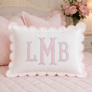 May include: A rectangular white throw pillow with a scalloped edge trimmed in light pink. The center features an embroidered monogram with the letters LMB, where the letters are filled with light pink thread and outlined in light blue thread.