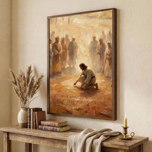 Puede incluir: Una impresión enmarcada representa una escena bíblica con figuras en tonos neutros. La figura central está arrodillada, escribiendo en el suelo, rodeada de otros. El texto "LET HIM WHO IS WITHOUT SIN CAST THE FIRST STONE" es visible. La obra de arte se exhibe sobre una mesa de madera.