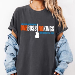 May include: Dark gray t-shirt with the text "ONE BOSS NO KINGS" in orange and white, with a white guitar graphic. The shirt also includes the text "LAND OF HOPE AND DREAMS TOUR" and "BOSTON MAY 24, 2026".