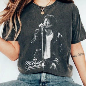 May include: Dark gray t-shirt featuring a black and white image of Michael Jackson performing on stage. The shirt has the text "Michael Jackson" in a cursive font. The shirt is a vintage style.