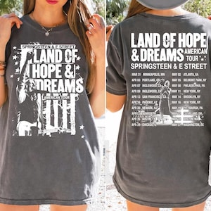 May include: Dark gray t-shirt featuring the text "LAND OF HOPE & DREAMS AMERICAN TOUR" with a graphic of a musician. The back of the shirt lists tour dates and locations for Springsteen & E Street.
