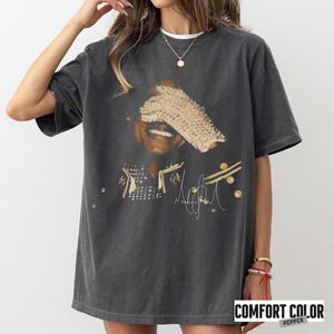 May include: Dark gray Comfort Colors t-shirt featuring a graphic of a smiling person with a gold-colored head covering. The shirt includes gold accents and a signature. The text "COMFORT COLOR PEPPER" is printed at the bottom.