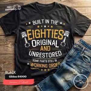 Op de afbeelding: Een zwart t-shirt met korte mouwen en goudkleurige en witte tekst met de tekst BUILT IN THE EIGHTIES ORIGINAL AND UNRESTORED SOME PARTS STILL IN WORKING ORDER, geflankeerd door twee illustraties van motorzuigers aan de linker- en rechterkant.