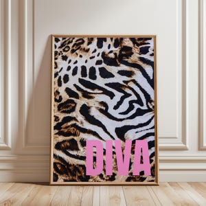 Op de afbeelding: Een ingelijste kunstdruk met een luipaard- en zebraprintpatroon in bruin, zwart en wit. Het woord "DIVA" wordt weergegeven in grote, roze letters. De lijst is lichtbruin van kleur. De print staat op een houten vloer.