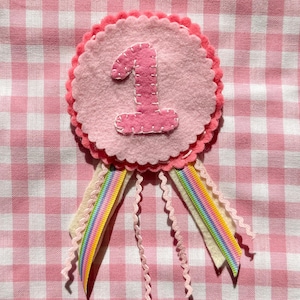 May include: A circular felt badge featuring a light pink number one stitched onto a pale pink scalloped base. The badge includes two cream-colored felt tails, two rainbow-striped ribbons, and three thin, wavy pink ribbons hanging downward.
