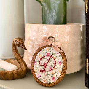 May include: An oval wooden embroidery hoop frame containing fabric with a floral pattern of small pink, yellow, and purple flowers. A dark red embroidered letter D is centered on the fabric. A small, light pink velvet ribbon bow is attached to the top.