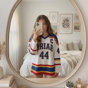 May include: White long-sleeved jersey with blue, red, and yellow stripes. The jersey features the word "BRIAR" and the number "44". The jersey has a V-neck collar. The jersey is worn by a person.