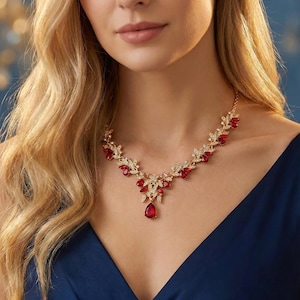 May include: A gold-toned necklace with red and clear gemstones. The necklace features a floral design with red teardrop-shaped gems and clear, round gems. The necklace is worn against a dark blue dress.