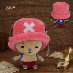 May include: A plush toy of a cartoon character, approximately 9.4 inches tall, with a pink hat featuring a white cross. The character has tan fur, a blue nose, and is wearing a blue and red outfit. A compass and map are in the background.