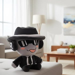 May include: A plush toy in the shape of a cartoon character, wearing a black fedora hat, sunglasses, and a black suit with a tie. The toy has gray skin, pink cheeks, and a smiling expression. The toy is sitting on a gray ottoman.