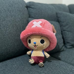 May include: A plush toy of a cartoon character with a pink hat featuring a white cross, brown ears, and a blue nose. The character has large black eyes, a small mouth, and is wearing a maroon outfit. The toy is sitting on a gray couch.