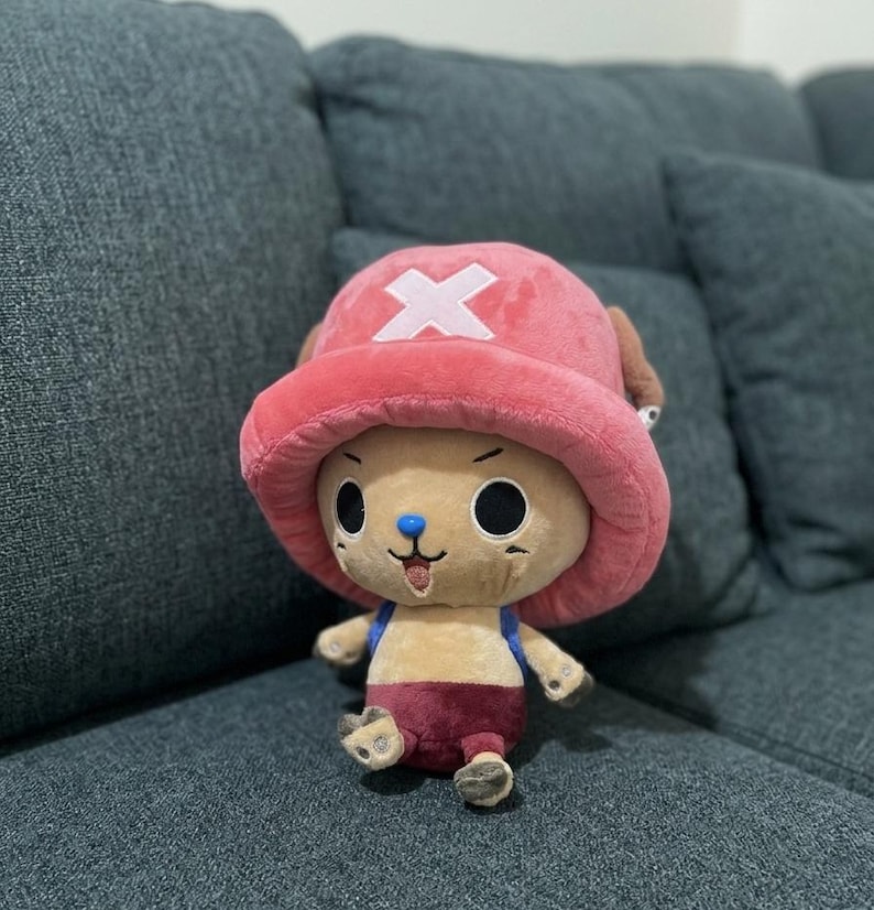 May include: A plush toy of a cartoon character with a pink hat featuring a white cross, brown ears, and a blue nose. The character has large black eyes, a small mouth, and is wearing a maroon outfit. The toy is sitting on a gray couch.