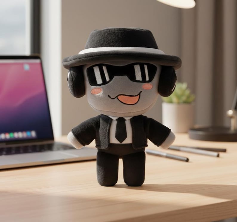 May include: A plush toy character wearing a black fedora hat, sunglasses, and a black suit with a tie. The character has a gray face with a smiling expression and pink cheeks. The toy is standing on a wooden desk.