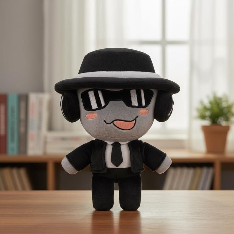 May include: A plush toy character wearing a black fedora hat with a white band, sunglasses, and a black suit with a tie. The toy has a gray face with a smiling expression and pink cheeks. The background includes a wooden table and a window.