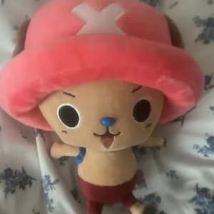 May include: A plush toy of a cartoon character with a large pink hat featuring a white cross. The character has a tan face, large black eyes, a blue nose, and a small open mouth. It is wearing red shorts and has small arms and legs.