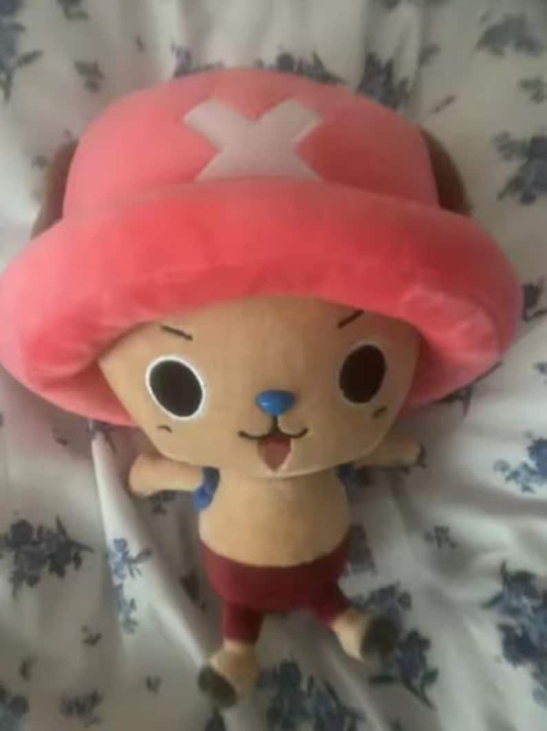 May include: A plush toy of a cartoon character with a large pink hat featuring a white cross. The character has a tan face, large black eyes, a blue nose, and a small open mouth. It is wearing red shorts and has small arms and legs.
