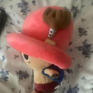 May include: A plush toy of a character with a large pink hat and brown antlers. The character has a tan body, red shorts, and a blue backpack. The toy is on a floral patterned surface.