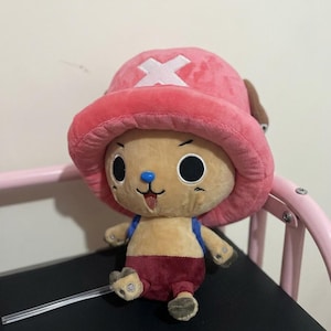 May include: A plush toy of a cartoon character with a pink hat featuring a white cross, a tan face, and a blue nose. The character wears a red outfit and has black eyes. The toy is sitting on a black surface.