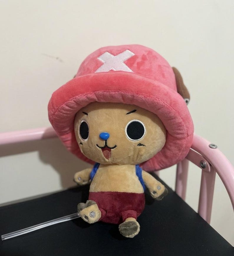 May include: A plush toy of a cartoon character with a pink hat featuring a white cross, a tan face, and a blue nose. The character wears a red outfit and has black eyes. The toy is sitting on a black surface.