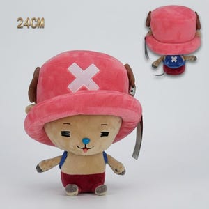 May include: A plush toy of a cartoon character, approximately 9.4 inches tall, with a pink hat featuring a white cross. The character has brown ears, a blue nose, and is wearing a blue vest and red shorts. A smaller version of the toy is also visible.