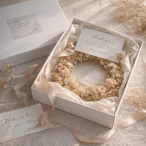 May include: A white box containing a floral wreath with pink roses and dried flowers, a "Thank you" card, and a card that says "Made with love for your wedding". Ribbons and tissue paper are also in the box.