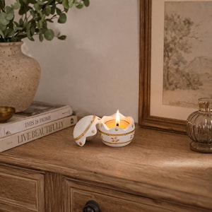 Może przedstawiać: Płonąca świeca w białym, jajowatym świeczniku ze złotymi kwiatowymi detalami. Świecznik jest otwarty, ukazując płonącą świecę. Stoi na drewnianej powierzchni, obok książek i wazonu z zielenią. Dekoracyjny przedmiot.