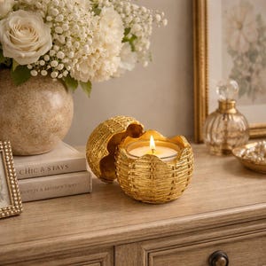 Może przedstawiać: Zapalone świeca w złotym pojemniku w stylu plecionym. Pojemnik jest otwarty, ukazując płomień świecy. Świeca stoi na drewnianej powierzchni, w tle wazon z białymi kwiatami.