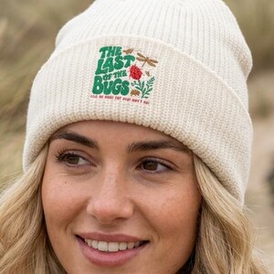 May include: A cream-colored ribbed knit beanie featuring a graphic print on the front. The design includes green text that reads THE LAST OF THE BUGS, accompanied by small illustrations of a dragonfly, a red flower, and various small insects.