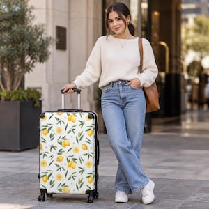 May include: A white rolling suitcase featuring a repeating pattern of yellow lemons, green leaves, and small white blossoms. The luggage has a black frame, black corner protectors, a retractable handle, and four black spinner wheels.