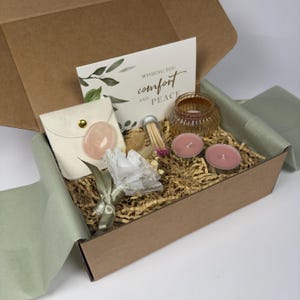 May include: A brown cardboard gift box containing a white card with the text Wishing You Comfort and Peace, a cream pouch with a pink stone, a small glass candle holder, two pink tea light candles, a glass vial of matches, and a white paper flower.