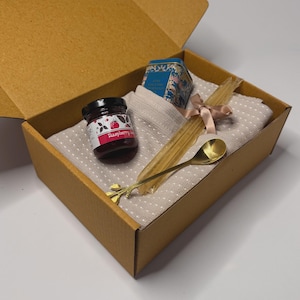 May include: A gift box containing raspberry jam, a small floral tin, a gold spoon, and straws. The items are arranged on a beige polka dot cloth, inside a brown cardboard box. The spoon is gold.