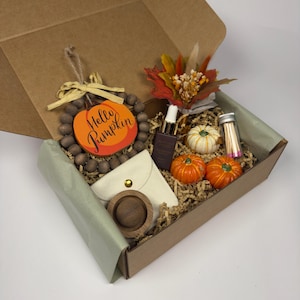 Peut inclure: Une boîte cadeau en carton contenant une couronne de perles en bois avec une pancarte citrouille portant l'inscription Hello Pumpkin, un petit bouquet de fleurs séchées, un flacon compte-gouttes en verre foncé, une pochette blanche et des allumettes.