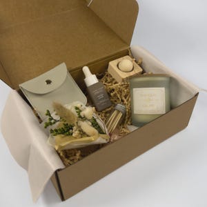 May include: A brown gift box containing self-care products. Inside are a candle, a small pouch, a serum bottle, a wooden block with a ball, a small dried flower bouquet, and a match jar. The candle label says "Tranquil, Calm Me."