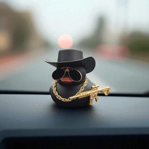 May include: A black rubber duck adorned with a black hat, gold-framed sunglasses, and a gold chain. It also features a gold toy gun. The duck is positioned on a car dashboard.