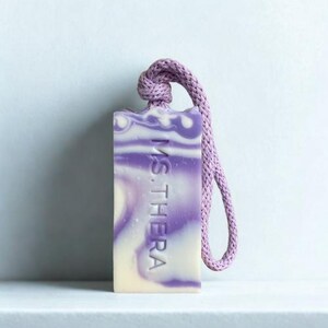 Puede incluir: Una pastilla de jabón rectangular con un diseño en espiral lavanda y blanco, sujeta a una cuerda lavanda. El jabón tiene el texto "MS. THERA" grabado. El jabón se exhibe en una estantería blanca.