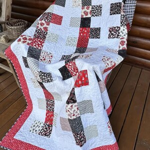 Könnte beinhalten: Eine Patchwork-Decke mit einem sich wiederholenden Muster aus weißen Quadraten, die sich mit schwarzen, roten und cremefarbenen Quadraten abwechseln, verziert mit weihnachtlichen Motiven wie Zuckerstangen, Weihnachtssterne und Tannenbäumen, mit rotem Blumenrand.