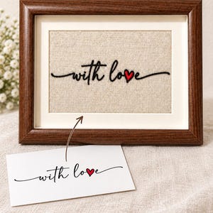 Puede incluir: Un marco de madera marrón oscuro que contiene un inserto de tela beige con el texto bordado en negro "with love" y un pequeño corazón rojo. Debajo del marco hay una tarjeta blanca con el mismo diseño de "with love" y corazón.