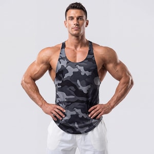 Puede incluir: Una camiseta sin mangas de camuflaje negra y gris. La parte superior atlética tiene un diseño de espalda de nadador y un dobladillo curvo. El patrón de camuflaje presenta tonos de gris y negro. La camiseta sin mangas está hecha de un material ligero y transpirable.