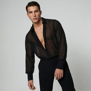 Può includere: Una camicia nera trasparente con bottoni, maniche lunghe e colletto classico. La camicia è abbinata a pantaloni neri. Il tessuto appare leggero e leggermente trasparente, creando un effetto a strati. La camicia è sbottonata sul petto.