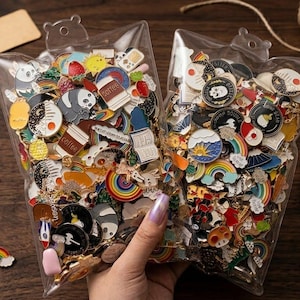 May include: Two clear plastic bags filled with colorful enamel pins. The pins feature various designs, including rainbows, coffee cups, pandas, and yin-yang symbols. The bags are held by a person with a light skin tone.