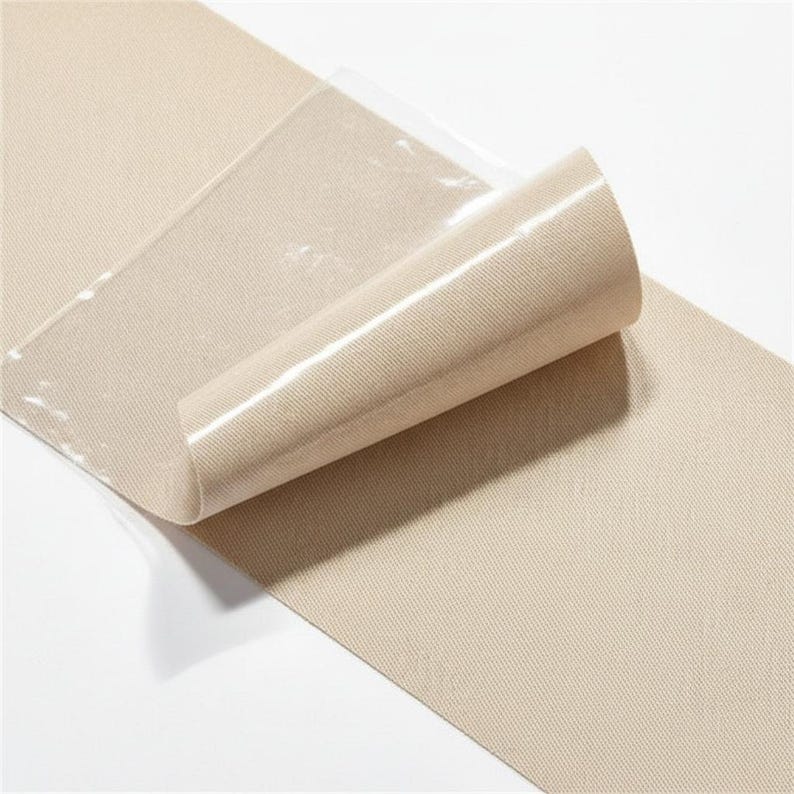 Pu&ograve; includere: Un rotolo di tessuto beige con uno strato protettivo trasparente parzialmente staccato. Il tessuto ha un aspetto strutturato e a coste. Lo strato trasparente &egrave; lucido e riflette la luce, rivelando il colore e la trama del tessuto.