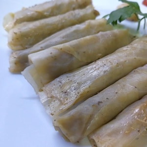 Può includere: Primo piano di diversi involtini di cavolo ripieni, disposti in fila su un piatto bianco. Gli involtini sono di colore beige chiaro, con macchie di condimento visibili. Un rametto di prezzemolo e un piccolo pomodoro sono visibili sullo sfondo.