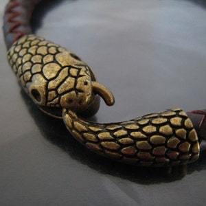 May include: A brown leather bracelet with a gold-toned snake head clasp. The snake head is detailed with scales and has a textured finish.